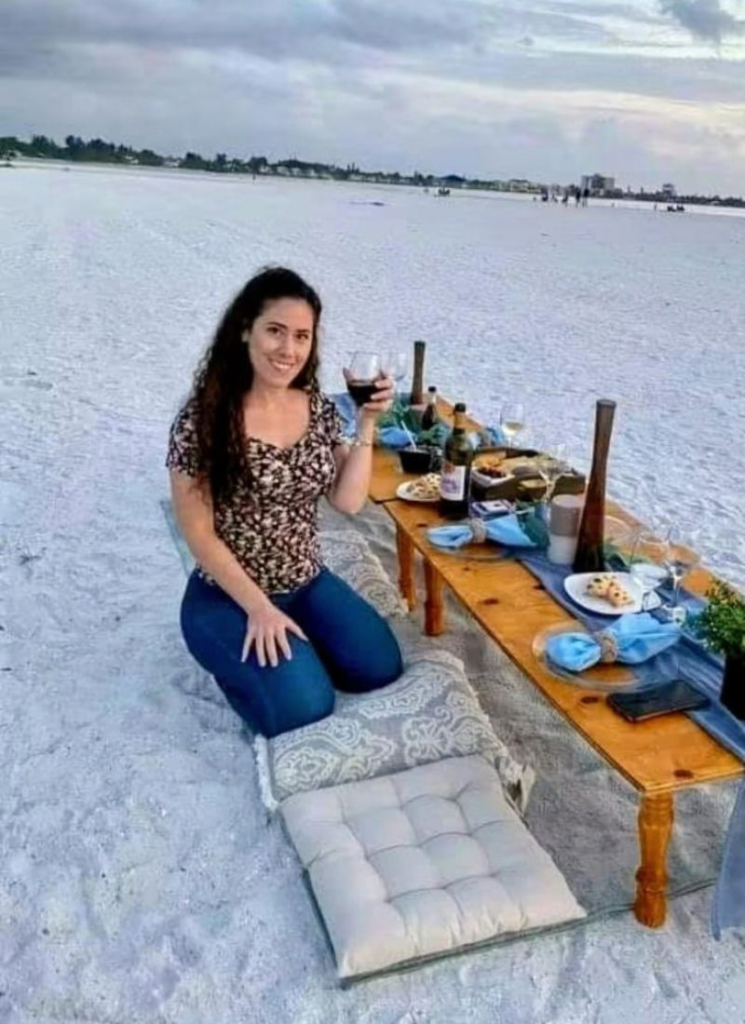Owner of Siesta Social Events Miranda Schlake enjoying a beautifully arranged beach picnic with a low wooden table, cushions and food by the water.