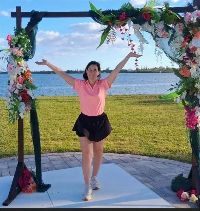 Costal outdoor event setting with colorful floral arrangement on wedding arch,blue water, and palm trees in background.