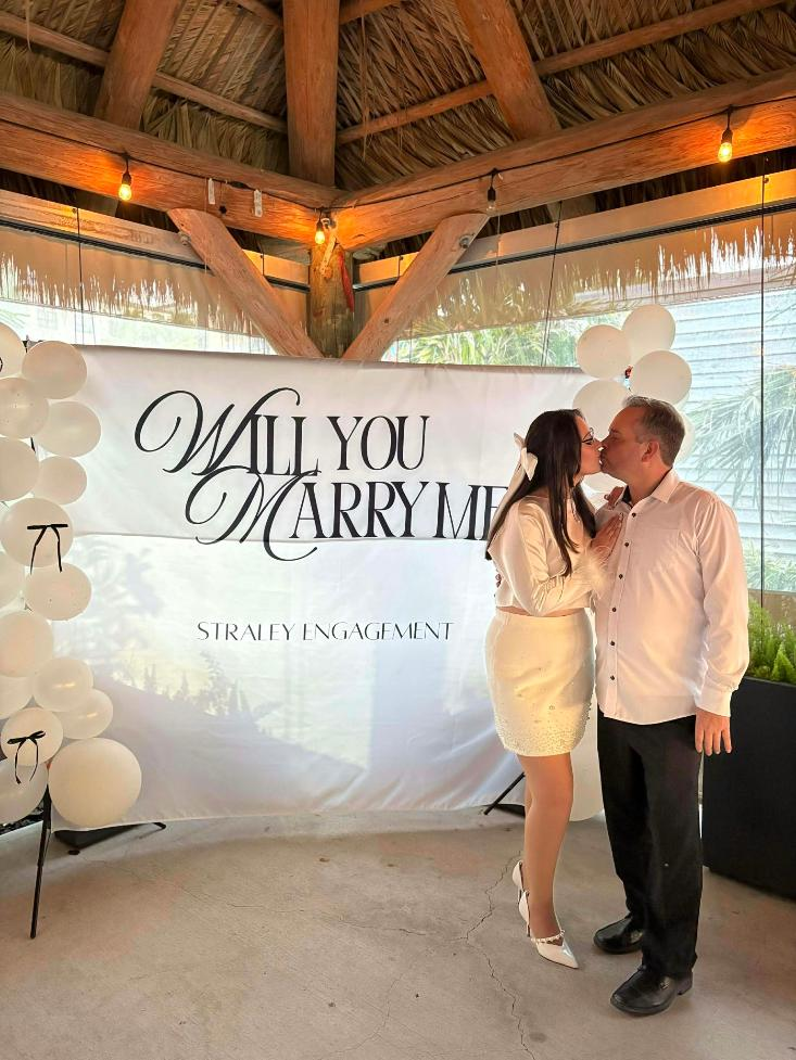 Couple kissing beneath a decorated pavillion beside a Will Yoiu Marry Me proposal backdrop.