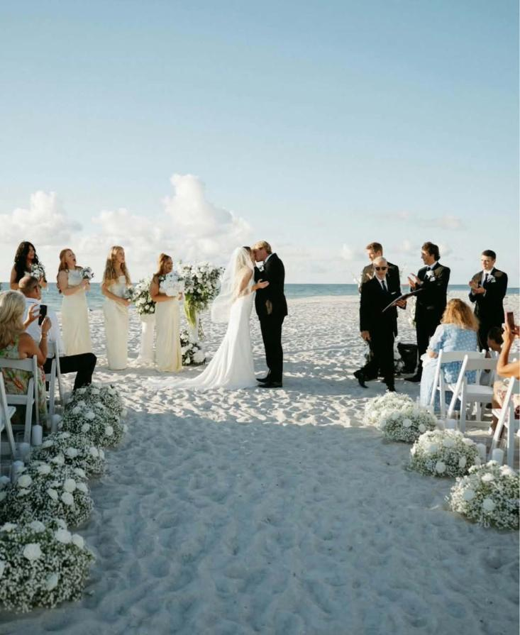 Beach wedding ceremony on the Gulf Coast with bride, groom, wedding party, and floral decorations along the sandy aisle.
