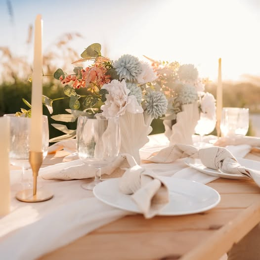 Elegant outdoor event table with white dinnerware, folded napkins, taper candles and soft floral centerpieces.