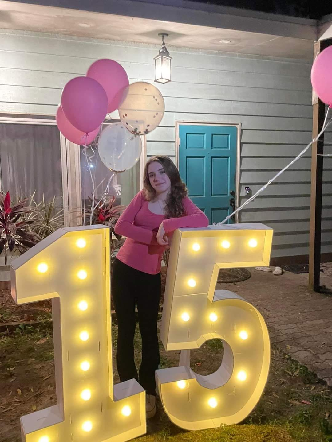 Teenage girl celebrating her fifteenth birthday beside illuminated number 15 decorations and pink balloons.